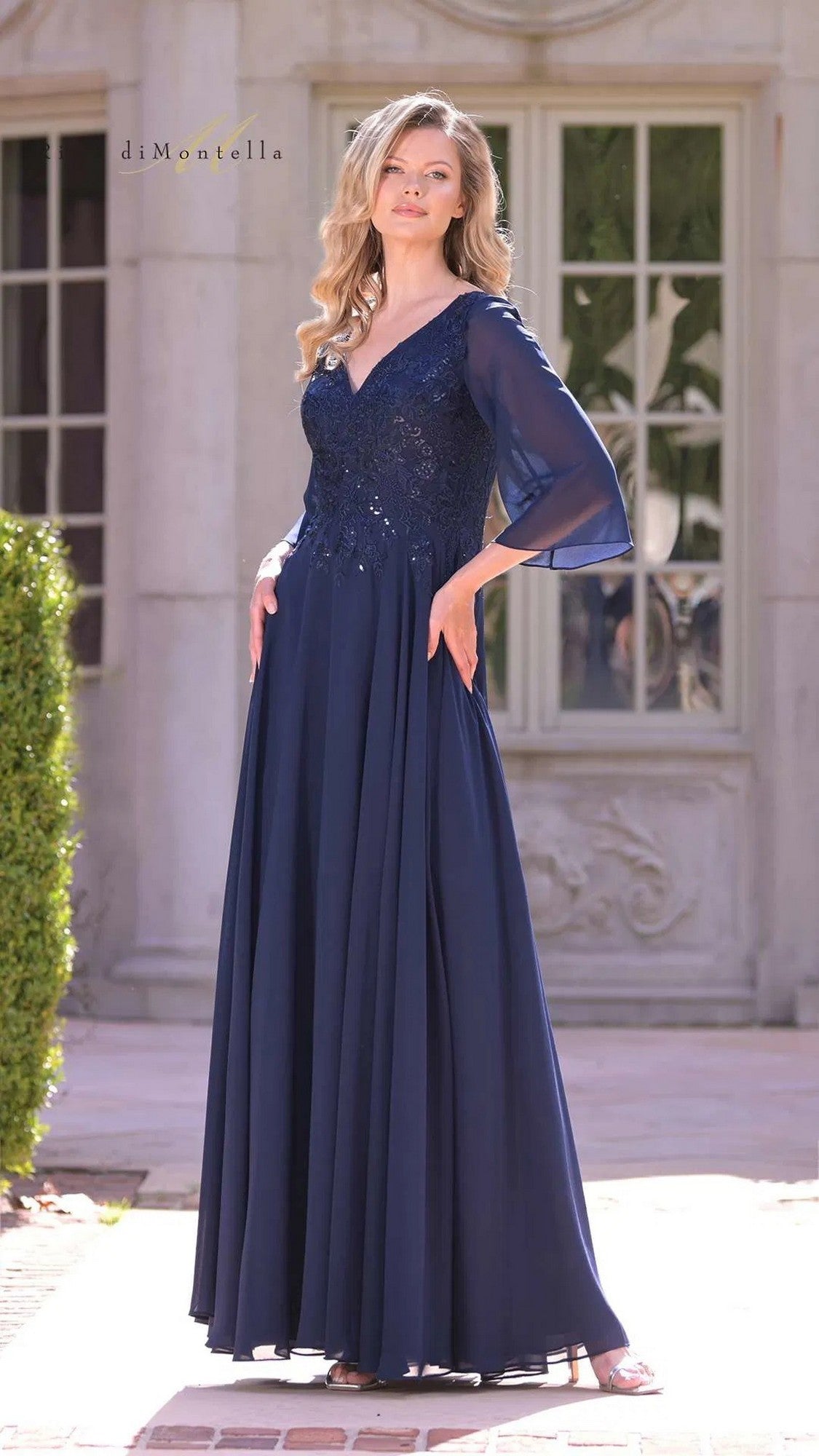 A woman stands outdoors wearing the Rina Di Montella RD3241 long formal dress in navy, featuring sheer sleeves and a beautifully embroidered floral bodice.